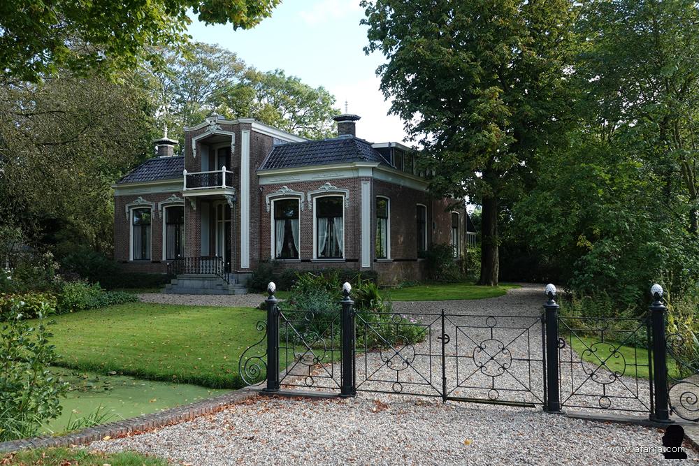 een groot huis achter een sloot en een mooi smeedwerkhek voor de oprit