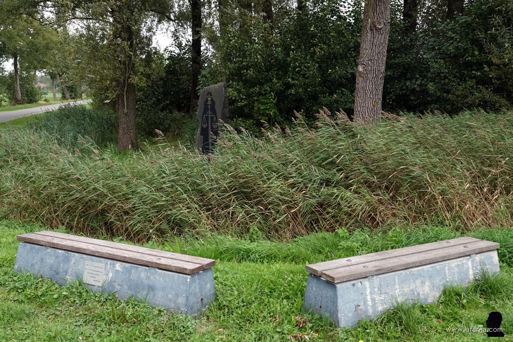 op de voorgrond twee bankjes, achter het riet staat op de achtergrond een beeld tussen de bomen