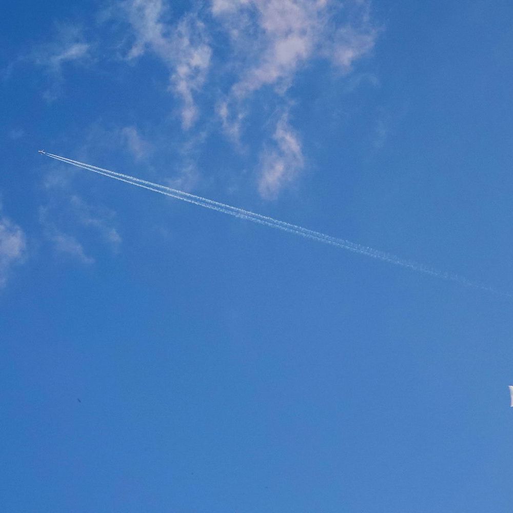 linksboven een straalvliegtuig tussen wolkenslierten en rechtsonder de maan