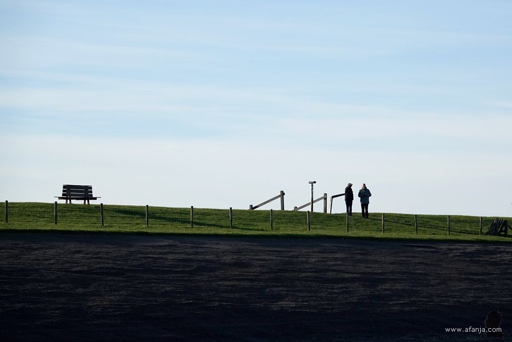 rechts staan twee mensen op de dijk, links staat een bankje