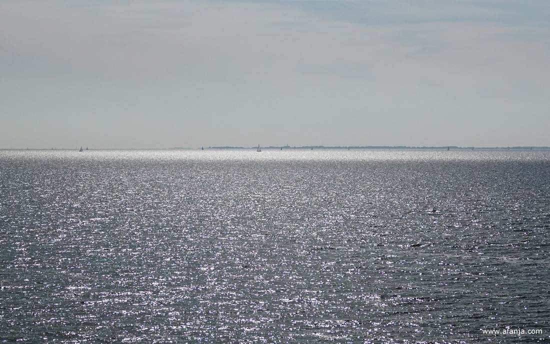 in de glinstering van het IJsselmeer zijn wat zeilbootjes te zien, daarachter ligt Noord-Holland