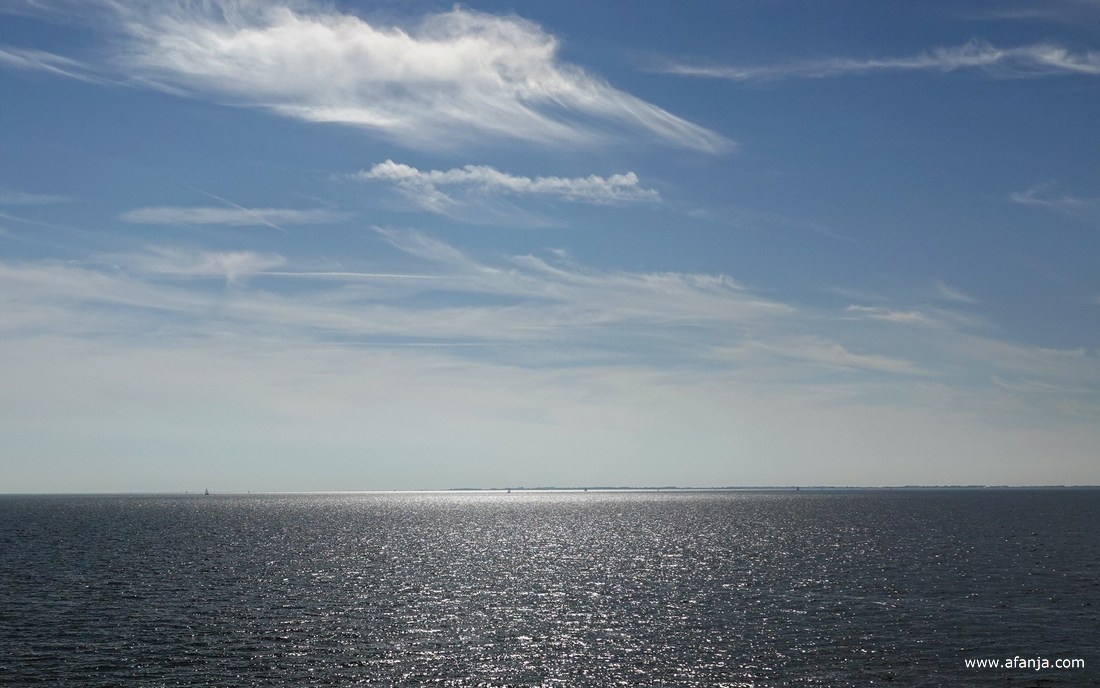 in de glinstering van het IJsselmeer zijn wat zeilbootjes te zien, daarachter ligt Noord-Holland