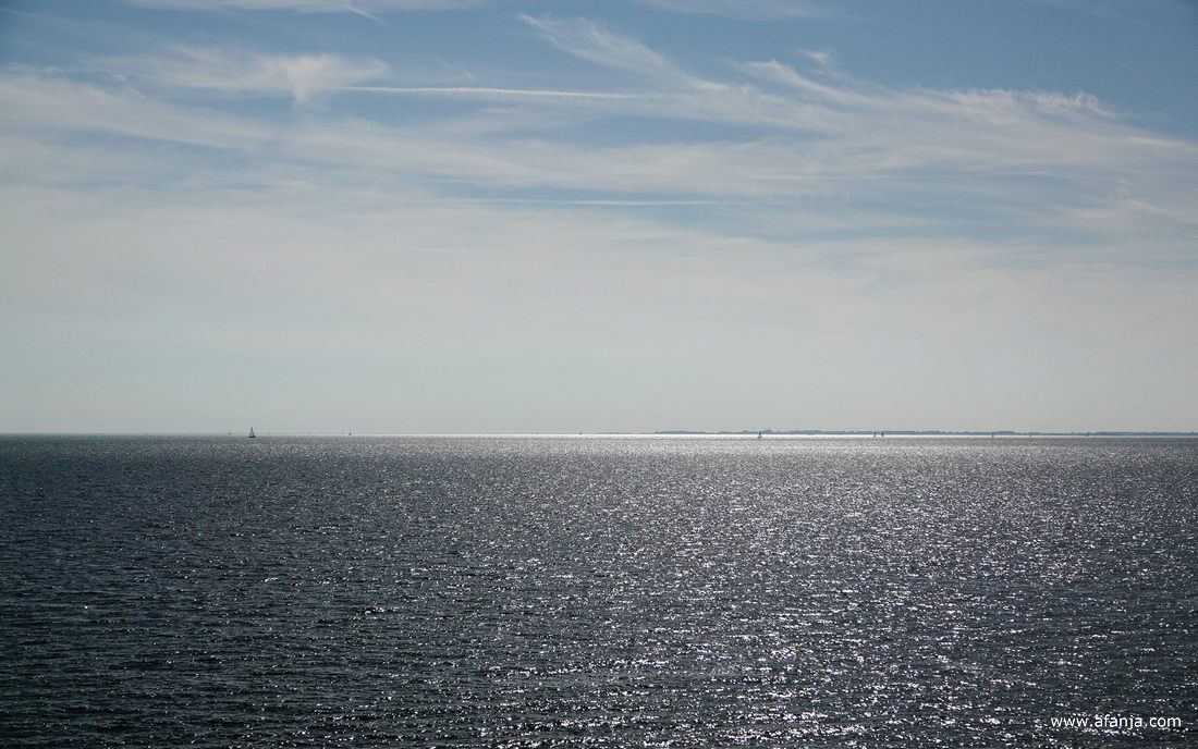 in de glinstering van het IJsselmeer zijn wat zeilbootjes te zien, daarachter ligt Noord-Holland
