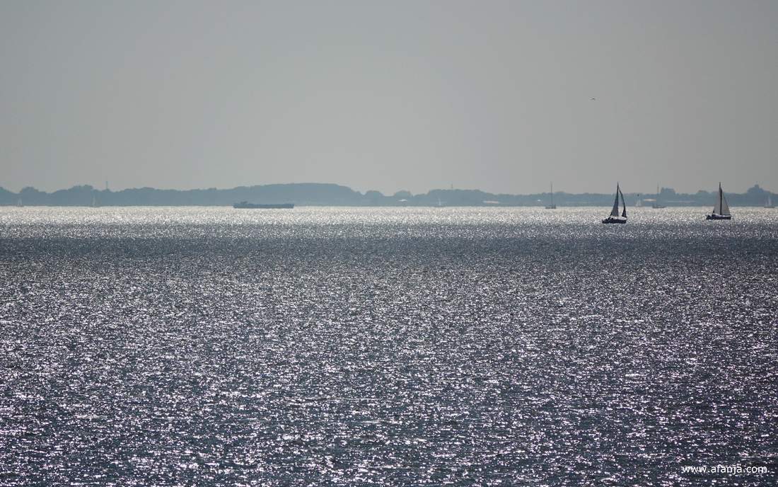 in de glinstering van het IJsselmeer zijn wat zeilbootjes te zien, daarachter ligt Noord-Holland
