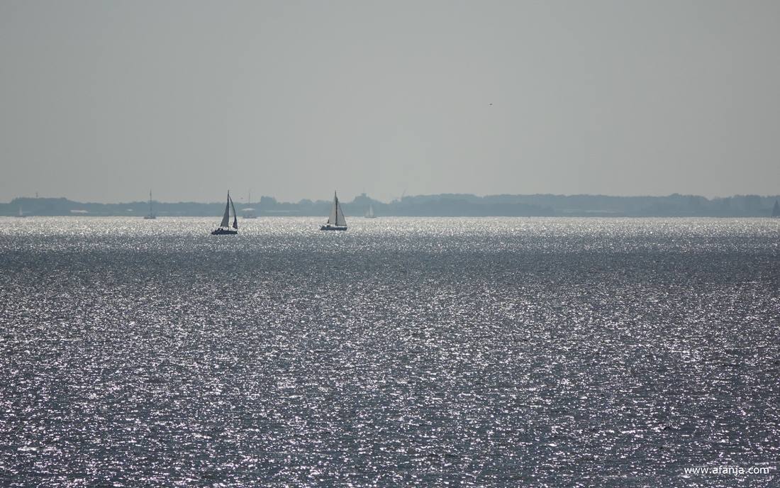 in de glinstering van het IJsselmeer zijn wat zeilbootjes te zien, daarachter ligt Noord-Holland